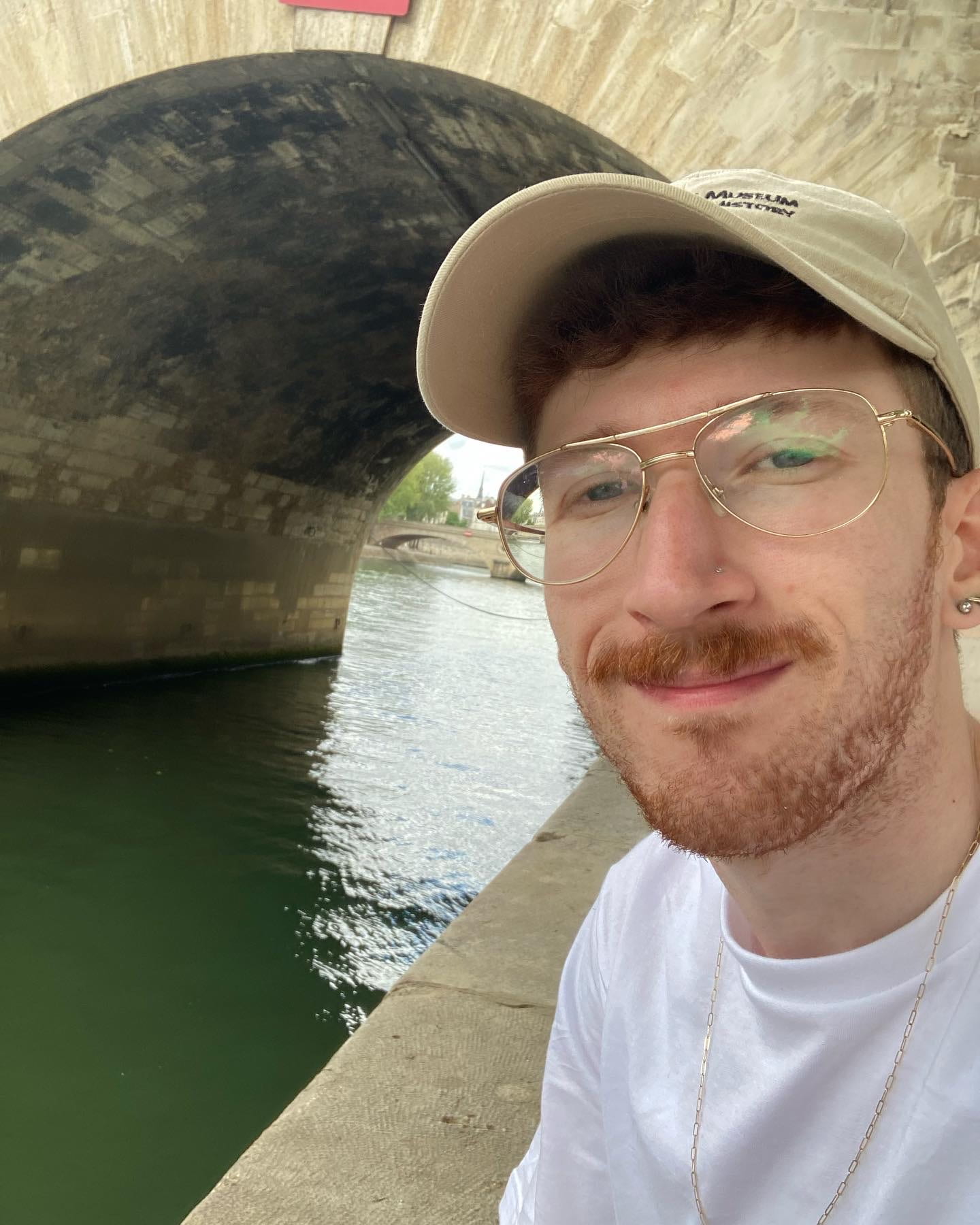 Young person in glasses, baseball cap, and baseball top posing by the Seine in Paris after visiting the Louvre.